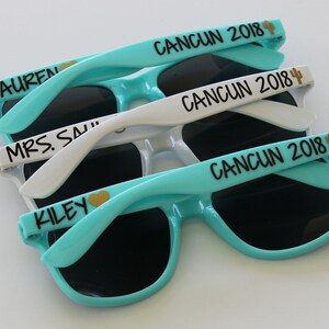 May include: Three pairs of sunglasses with black frames and dark lenses. The sunglasses are stacked on top of each other. The top pair is white with the text "CANCUN 2018" and a cactus icon. The middle pair is white with the text "CANCUN 2018" and a cactus icon. The bottom pair is turquoise with the text "CANCUN 2018" and a cactus icon.