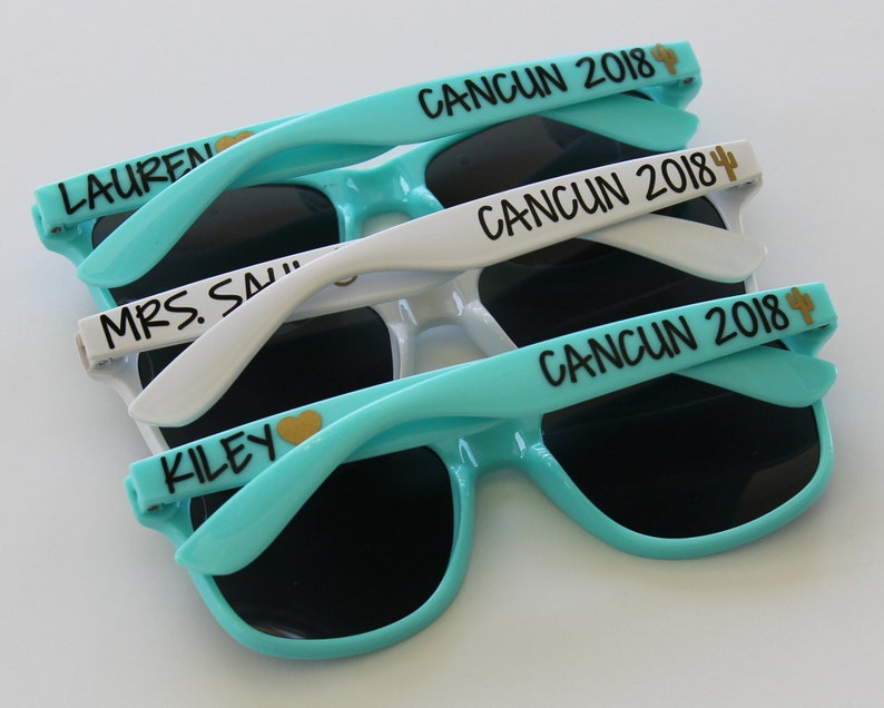 May include: Three pairs of sunglasses with black frames and dark lenses. The sunglasses are stacked on top of each other. The top pair is white with the text "CANCUN 2018" and a cactus icon. The middle pair is white with the text "CANCUN 2018" and a cactus icon. The bottom pair is turquoise with the text "CANCUN 2018" and a cactus icon.