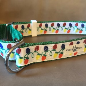 May include: A green and white dog collar featuring a repeating pattern of colorful Christmas light bulbs. The collar includes two silver metal D-rings, a white plastic slider, a red buckle, and a white fabric tag labeled with Koda's Collars.
