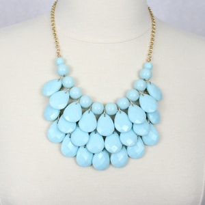 May include: A light blue statement necklace with a cascading teardrop design. The necklace is made of plastic beads and has a gold chain.