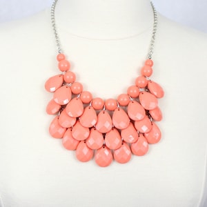 May include: A coral-colored statement necklace with a cascading design of teardrop-shaped beads. The necklace is made of plastic beads and has a silver chain.