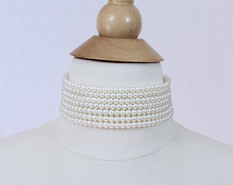 May include: A white faux pearl choker necklace with multiple strands of pearls. The necklace is displayed on a white mannequin bust.