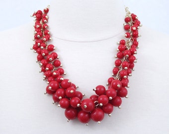 Puede incluir: Un collar de cuentas rojas con un diseño de racimo. El collar presenta numerosas cuentas rojas redondas de varios tamaños, ensartadas con una cadena dorada. Las cuentas están agrupadas para crear una pieza llamativa.