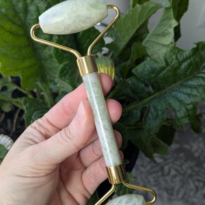 May include: A jade facial roller with a gold-colored metal frame. The roller has two rounded jade stones and a cylindrical jade handle. The background features green foliage.