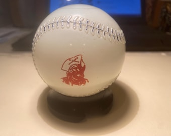 May include: A white baseball with red stitching and a red graphic of a pirate. The baseball is sitting on a surface, casting a shadow. The baseball is a sports collectible.