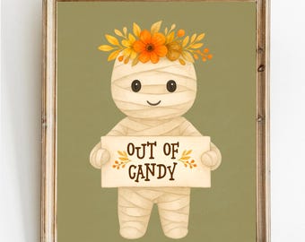 May include: A framed Halloween-themed print featuring a mummy character with a floral crown. The mummy holds a sign that reads "OUT OF CANDY" in a decorative font. The background is a muted green, and the frame is a light brown.