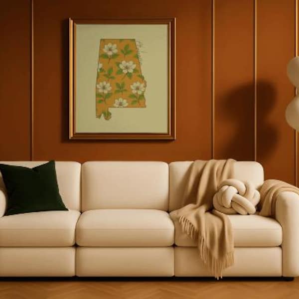 May include: A cream-colored sofa with a dark green pillow, a tan throw blanket, and a decorative knot pillow. A framed print of the state of Alabama with a floral pattern hangs on the brown wall. A white lamp stands to the right.