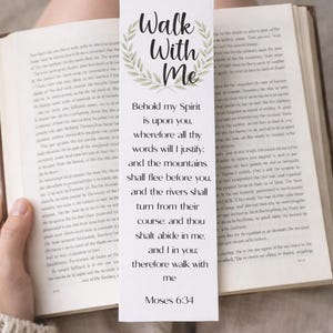 May include: A white bookmark with the text "Walk With Me" and a floral wreath design. The bookmark features a Bible verse from Moses 6:34. The bookmark is placed in an open book.