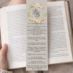 May include: A bookmark with a floral wreath design and the initials "RS". The bookmark has text about faith and sisterhood. The bookmark is held open in a book.