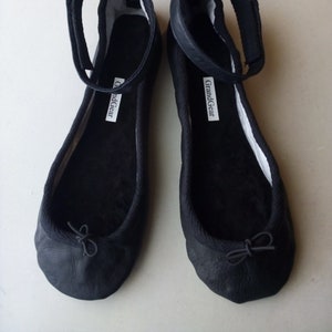 Könnte beinhalten: Ein Paar schwarze Ballerinas mit Knöchelriemen. Die Schuhe haben eine runde Spitze, eine kleine Schleife und ein weißes Etikett mit dem Text "GrandGear". Die Ballerinas sind aus einem weichen, strukturierten Material.