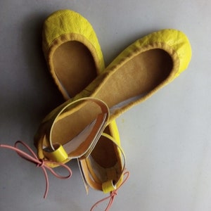 May include: A pair of yellow leather ballet flats with pink ribbon ties. The flats have a simple design and are made of soft leather.