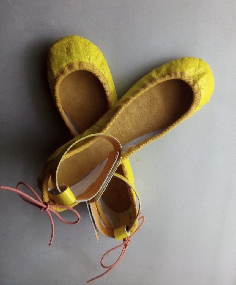 May include: A pair of yellow leather ballet flats with pink ribbon ties. The flats have a simple design and are made of soft leather.