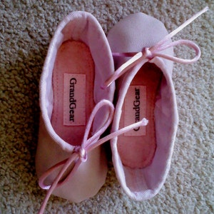 May include: A pair of pink ballet slippers with satin ribbons. The slippers have the brand name "GrandGear" printed on the inside.
