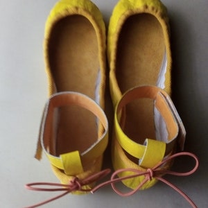 May include: A pair of yellow ballet flats with a pink ribbon tied around the ankles. The flats have a yellow strap across the top of the foot.