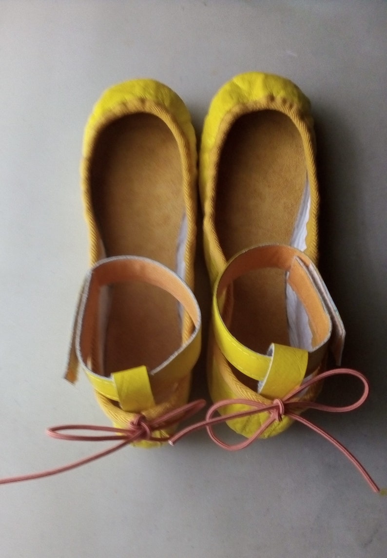 May include: A pair of yellow ballet flats with a pink ribbon tied around the ankles. The flats have a yellow strap across the top of the foot.