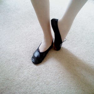 May include: A pair of black leather ballet shoes with elastic straps. The shoes are being worn by a person.