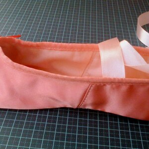 May include: A pink satin ballet slipper with white satin ribbons tied around the ankle.