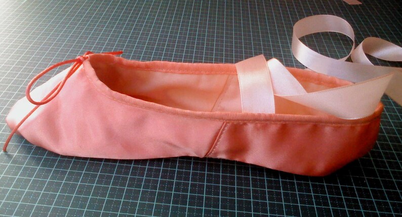 May include: A pink satin ballet slipper with white satin ribbons tied around the ankle.
