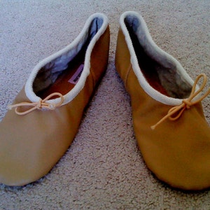 May include: A pair of tan leather ballet slippers with white fabric lining and a light pink ribbon tied at the top.