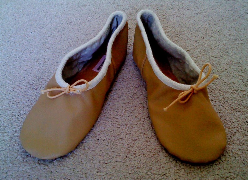 May include: A pair of tan leather ballet slippers with white fabric lining and a light pink ribbon tied at the top.