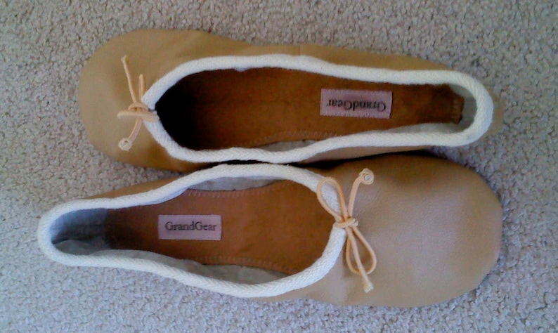 May include: A pair of tan leather ballet flats with white trim and a bow on the toe. The flats have the brand name "GrandGear" printed on the inside.