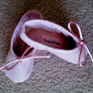 May include: A pair of pink ballet slippers with a ribbon tie. The slippers have the brand name "GrandGear" printed on the top.