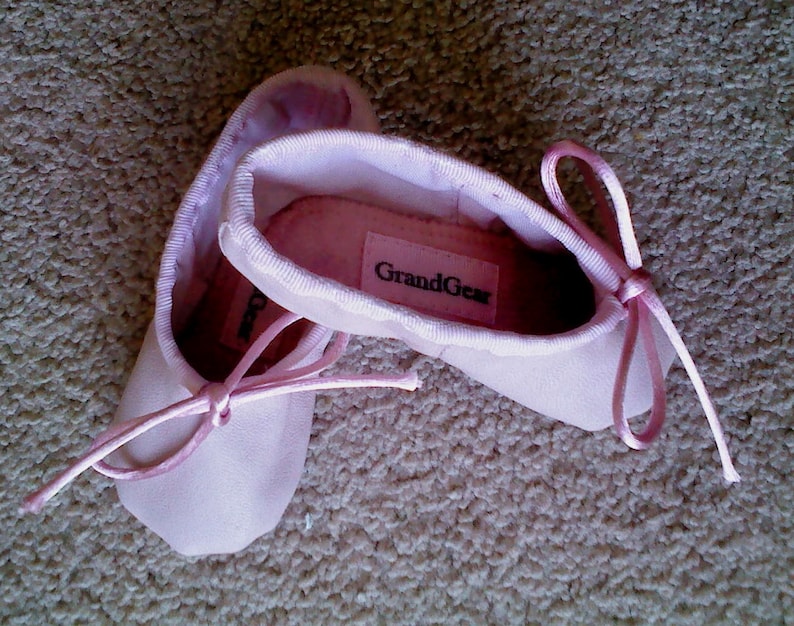 May include: A pair of pink ballet slippers with a ribbon tie. The slippers have the brand name "GrandGear" printed on the top.