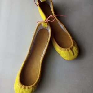 May include: A pair of yellow ballet flats with pink ribbon ties. The shoes are made of a soft, suede-like material.