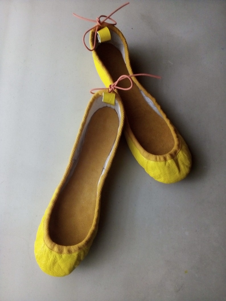 May include: A pair of yellow ballet flats with pink ribbon ties. The shoes are made of a soft, suede-like material.