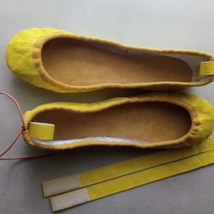 May include: A pair of yellow ballet flats with a light brown lining and a pair of yellow straps with white stitching.