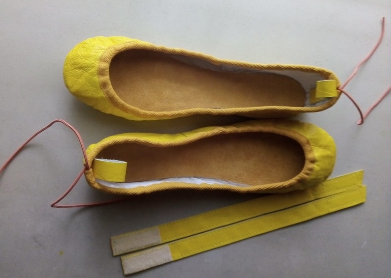 May include: A pair of yellow ballet flats with a light brown lining and a pair of yellow straps with white stitching.