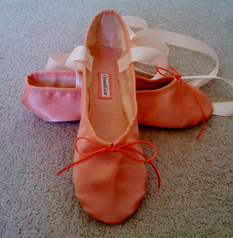May include: A pair of peach-colored satin ballet shoes with white ribbon ties and orange satin bows. The shoes have a label that reads "Grand Gear".