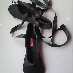 Op de afbeelding: Een paar zwarte satijnen ballerina's met zwarte satijnen linten. De ballerina's hebben een roze label met de tekst "Grand Gear".