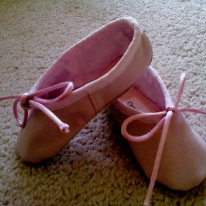 May include: A pair of pink ballet shoes with satin ribbons. The shoes are tied with a bow and are sitting on a light beige carpet.