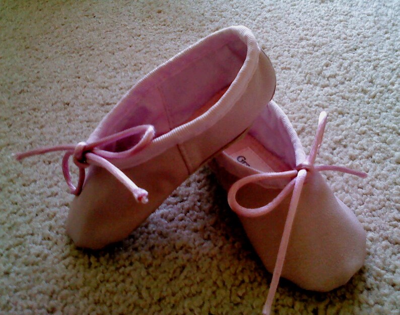 May include: A pair of pink ballet shoes with satin ribbons. The shoes are tied with a bow and are sitting on a light beige carpet.
