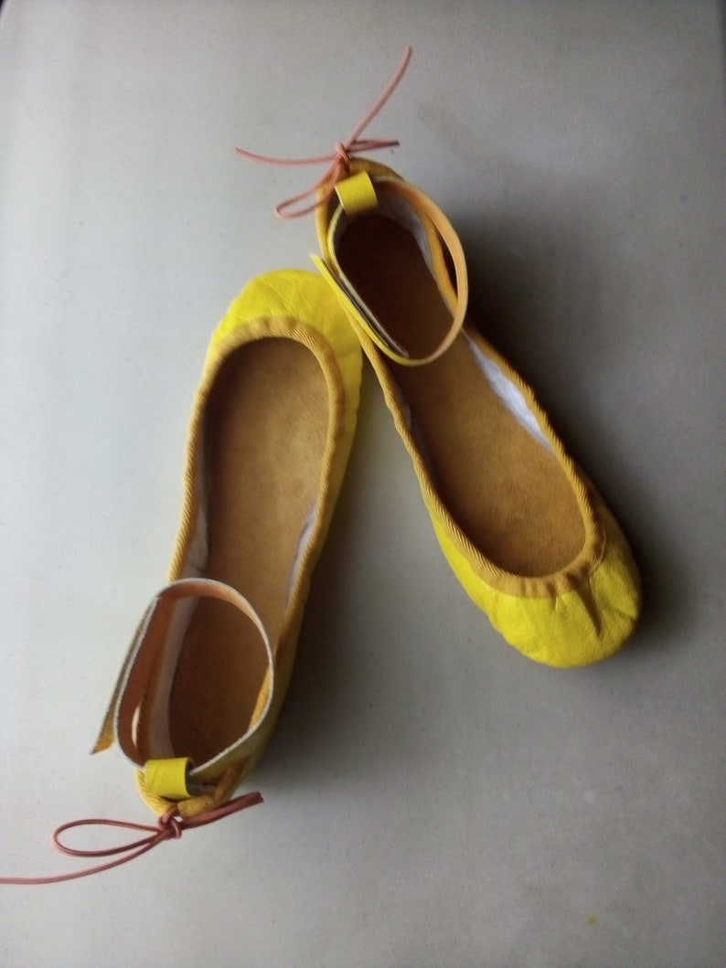 May include: A pair of yellow ballet flats with ankle straps and pink laces. The flats are made of a soft, suede-like material.