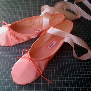 May include: A pair of pink satin ballet shoes with pink ribbon ties. The shoes are on a green gridded surface. The label on one shoe reads "Crandall-Cesar".