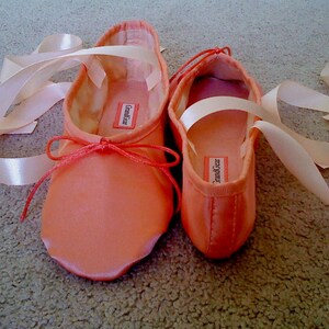 May include: A pair of pink satin ballet shoes with white satin ribbons. The shoes have the brand name "Grand Gear" printed on the inside.
