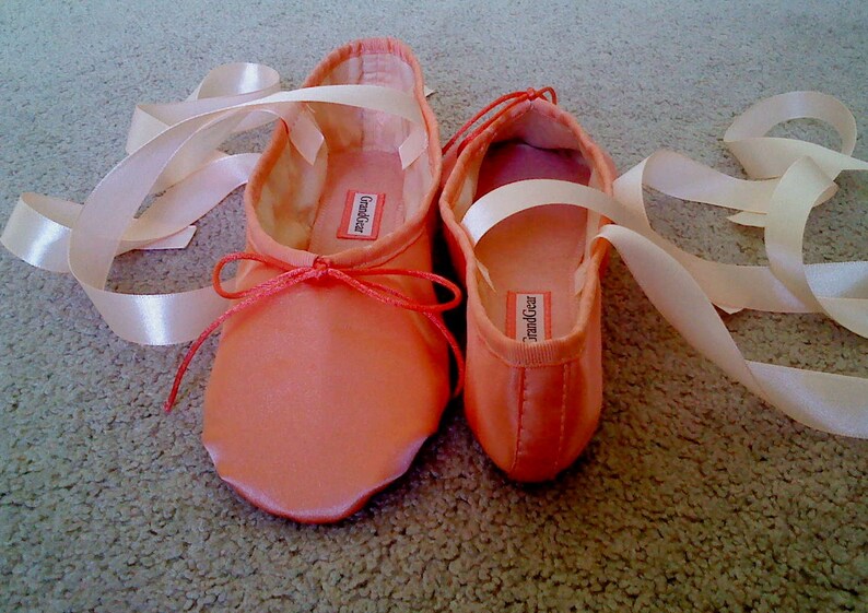 May include: A pair of pink satin ballet shoes with white satin ribbons. The shoes have the brand name "Grand Gear" printed on the inside.