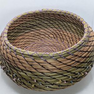 May include: Handwoven basket with a spiral design. The basket features a mix of natural colors, including green, brown, and tan, with a lavender-colored thread around the rim. The basket is adorned with small, colorful beads.