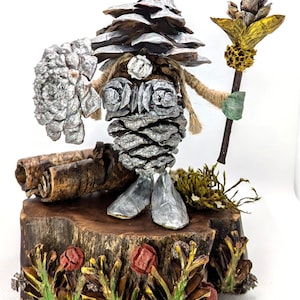 May include: A whimsical sculpture of a forest gnome crafted from pine cones, painted silver and brown. The gnome holds a staff and a silver-painted pine cone. It stands on a wooden base with painted foliage and red flowers.