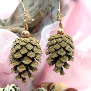 Puede incluir: Pendientes hechos a mano con colgantes de piña. Los pendientes tienen ganchos dorados y detalles de alambre de color cobre. Cada piña es de color verde-marrón apagado, con una pequeña cuenta dorada en la parte superior.