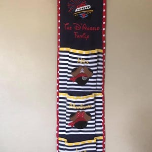 May include: A blue and white striped fabric wall organizer with red polka dots. It has four pockets with Mickey Mouse ears and pirate hats. The top pocket has a red, white, and blue cruise ship with fireworks and the text "Yo Ho Yo Ho The D'Angelo Family".