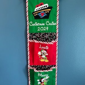 May include: A green, red, and black fabric Christmas stocking holder with a Mickey Mouse design. The top section features a ship with the text "Christmas Cruise 2024". The bottom section has two pockets with the names "Leslie" and "Mark" embroidered on them.
