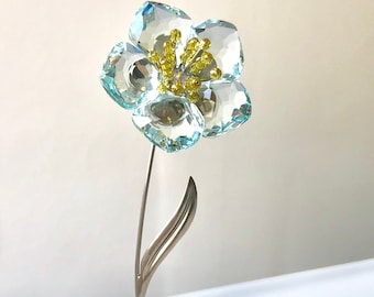 May include: A crystal flower sculpture with a light blue flower and a gold stem and base.