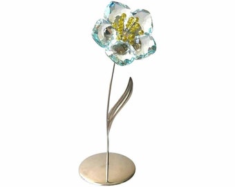 May include: A crystal flower figurine with a light blue flower and a yellow center. The flower is on a silver stand with a round base.