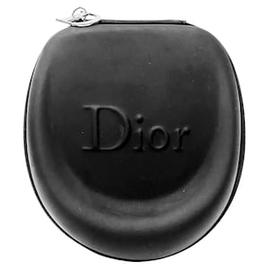 May include: A black, semi-rigid, rounded case with a zipper closure and a silver-toned metal charm attached to the zipper pull. The word Dior is embossed in the center of the front surface. The case has a smooth, matte texture and a compact shape.