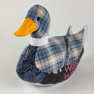 Puede incluir: Un pato de tela con cuerpo a cuadros azules, detalles de cachemira y pico naranja. El pato tiene una banda para el cuello a rayas blancas y grises y las palabras "WISH" y "Wishes" en rosa en su base. Un artículo decorativo.