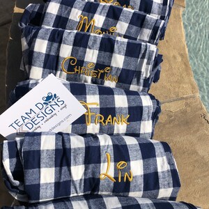 May include: A stack of seven folded navy blue and white buffalo plaid blankets, each featuring a name embroidered in gold thread. A white rectangular card with the text TEAM DOG DESIGNS and a frog logo is placed on the fourth blanket from the top.