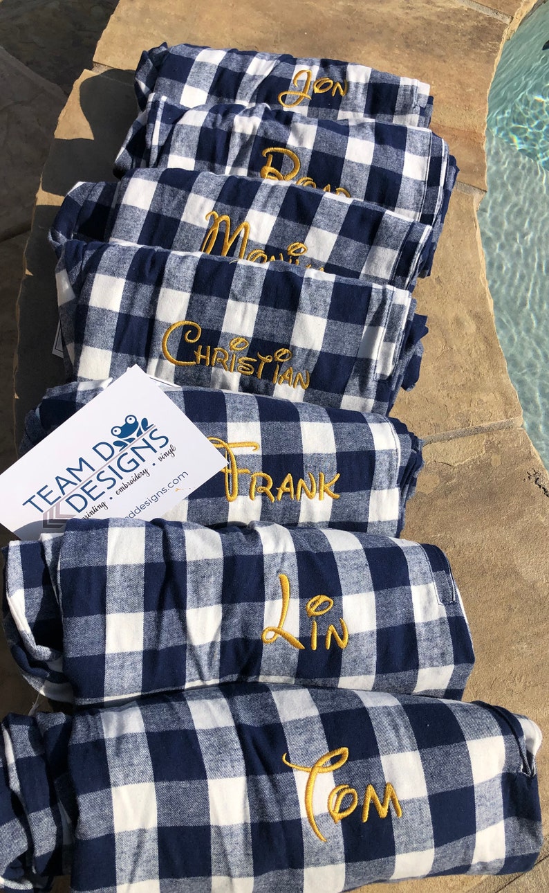 May include: A stack of seven folded navy blue and white buffalo plaid blankets, each featuring a name embroidered in gold thread. A white rectangular card with the text TEAM DOG DESIGNS and a frog logo is placed on the fourth blanket from the top.